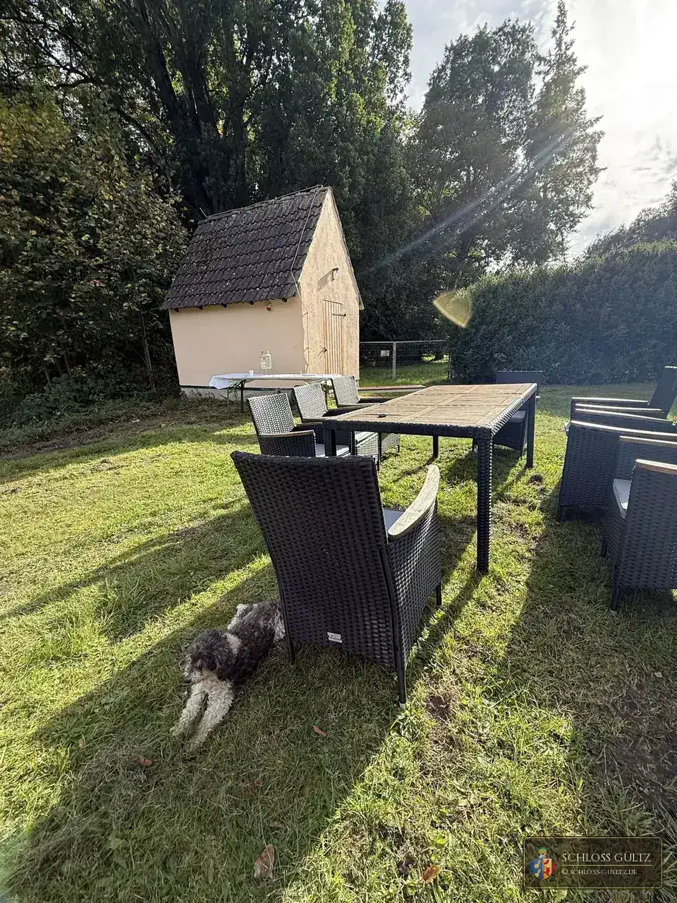 Sitzgruppe aus dunklen Geflechtsesseln um einen Holztisch auf dem Rasen vor dem hell verputzten Waschhaus mit Holztür; davor liegt ein grau-weißer Hund im Schatten