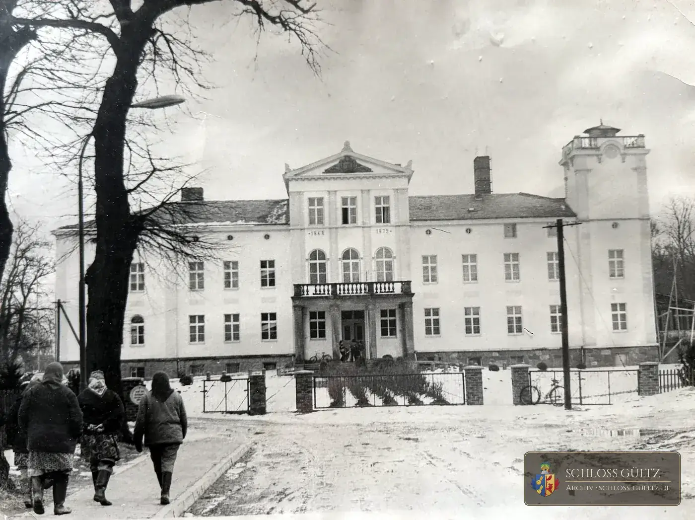 Schloss Gültz im Winter zur DDR-Zeit: Hofseite mit Mittelrisalit, Baudaten 1868 und 1872, Balkon über dem Portikus, Turm mit Aufsatz; im Vordergrund drei Frauen in Wintermänteln, Straßenlaterne, Zaun