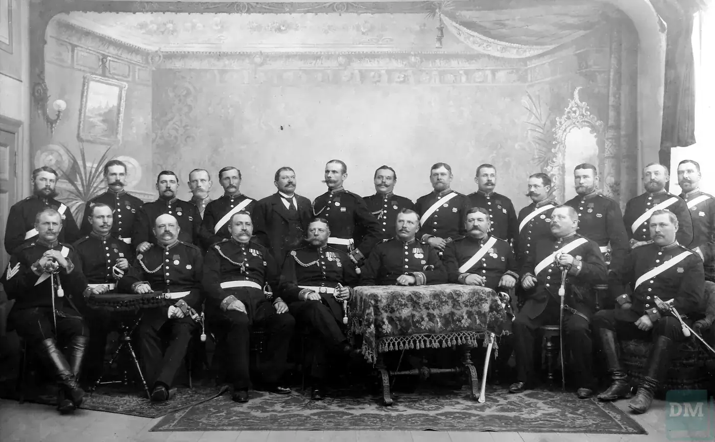 Historische Atelieraufnahme: rund zwanzig Männer in dunklen Ulanen-Uniformen mit weißem Bandelier, Säbeln und Orden, sitzend und stehend vor gemalter Salonkulisse, einer in Zivil