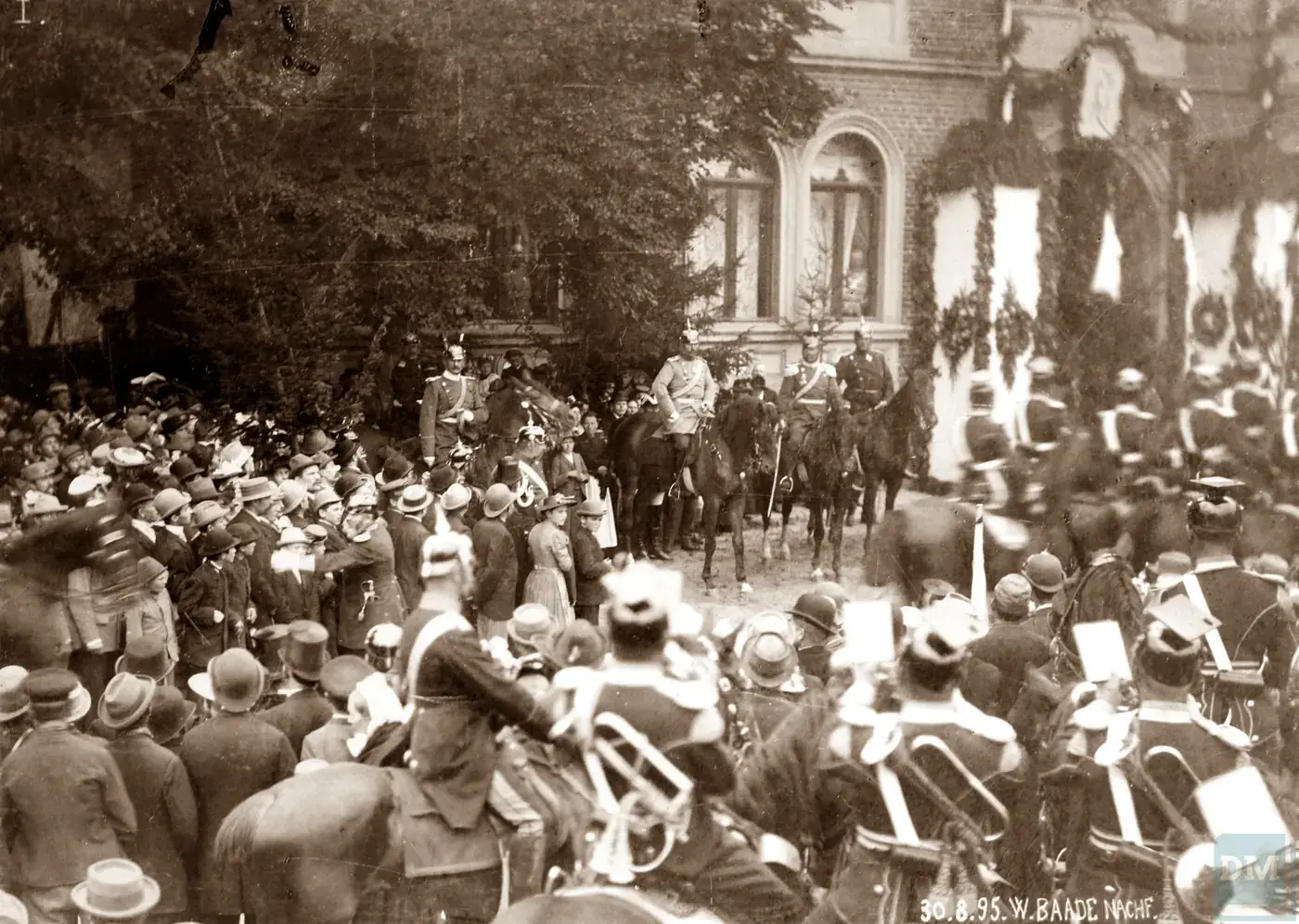 Historisches Foto: Kaiser Wilhelm II. zu Pferd mit Offizieren vor dem geschmückten Offizierkasino in Demmin, davor dichtgedrängte Zuschauer und vorbeireitende Ulanen mit Trompeten